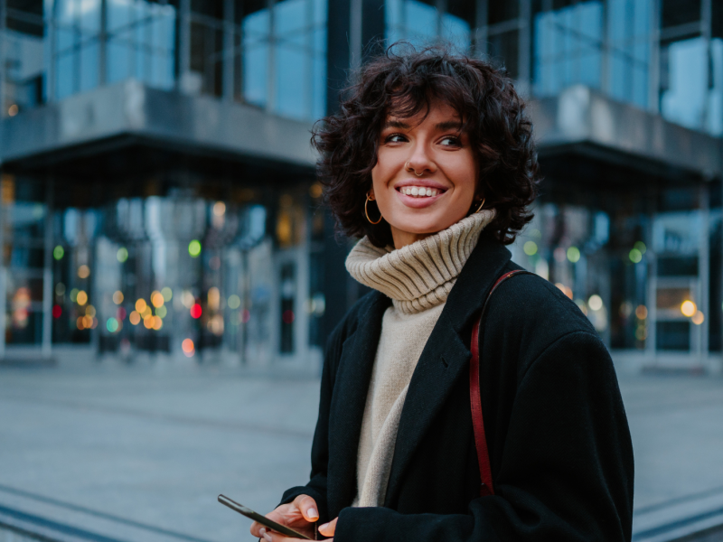 Frau vor einem Gebäude mit Handy in der Hand, die zur Seite lächelt