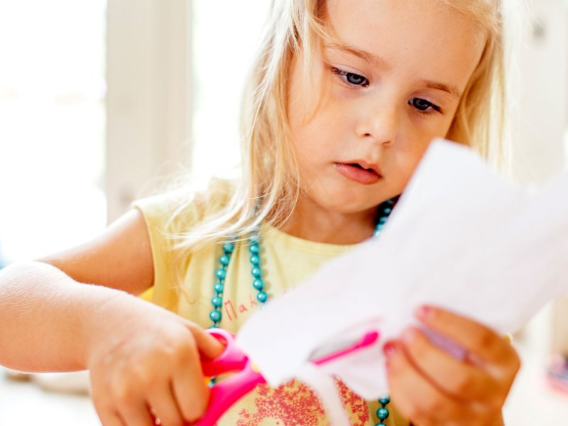 Mädchen im Vorschulalter schneidet konzentriert mit einer Schere ein Stück weißes Papier.