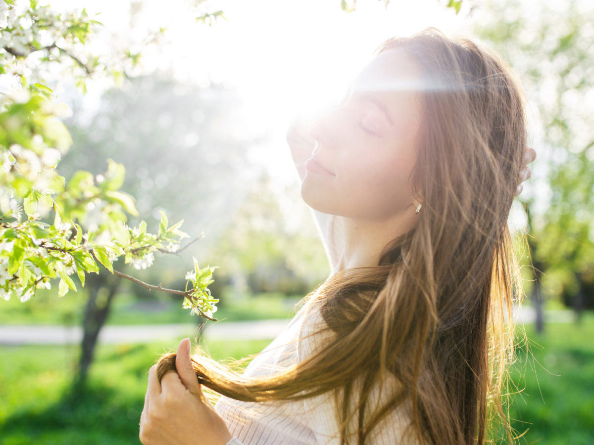 Frau, die in einem Park vor einem Baum steht und von einem Sonnenstrahl beleuchtet wird, während sie sich durch die Haare geht