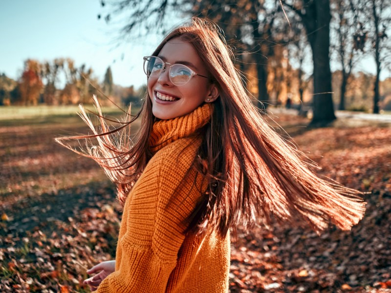 Frau im Herbst mit Brille und oranagenem Pulli, die sich lachend Richtung Sonne dreht