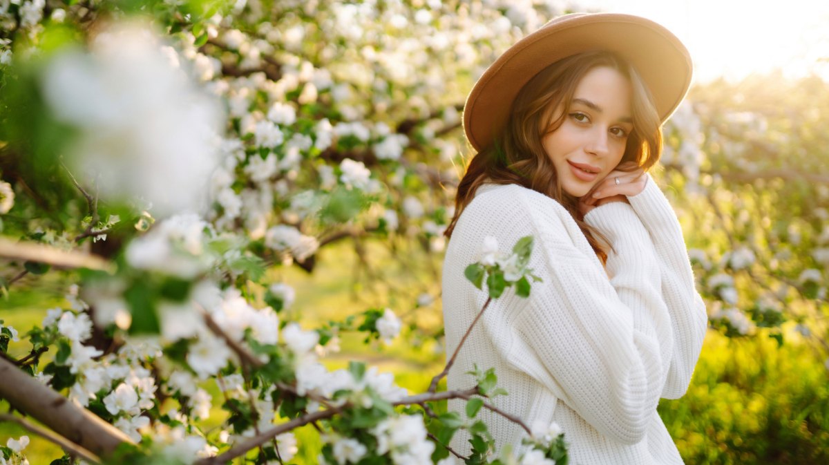 Frau mit Pullover und Hut steht vor einem blühenden Baum