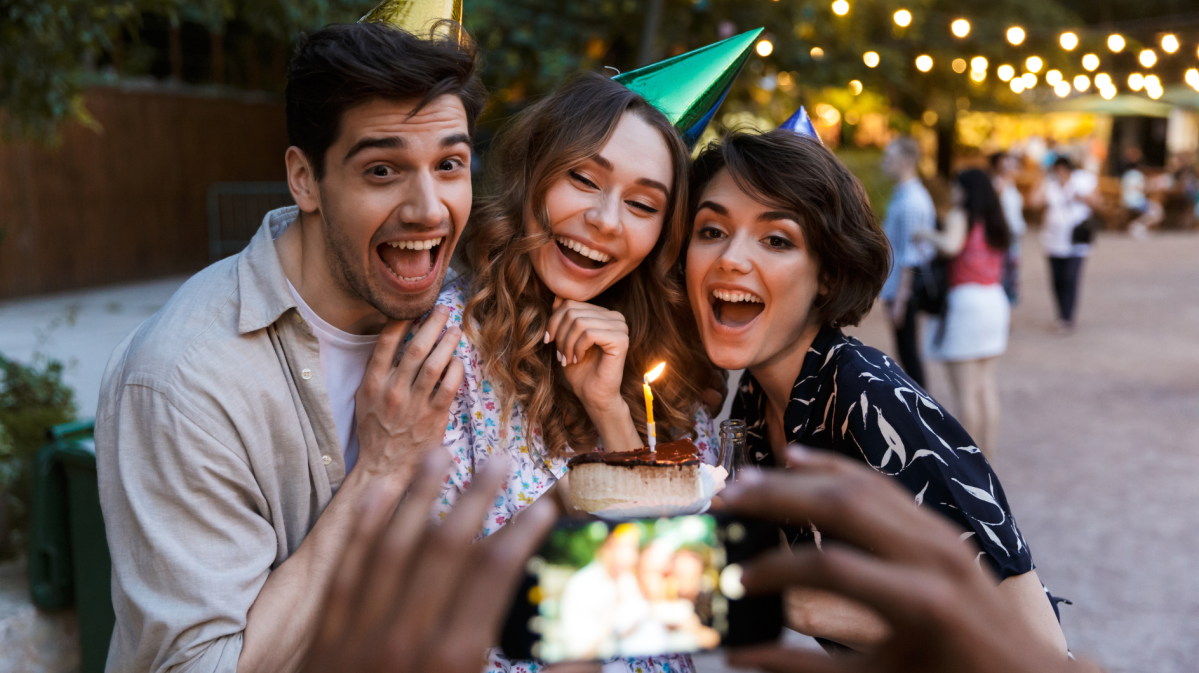 Zwei Frauen und ein Mann mit Partyhut und Kuchen in der Hand, von denen ein Foto gemacht wird