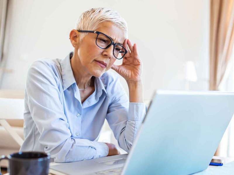 Ältere Frau mit kurzen weißen Haaren sitzt nachdenklich vor ihrem Laptop.
