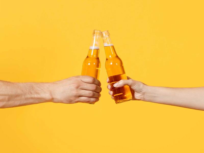 Close up beschnitten von Frau und Mann zwei Hände horizontal halten Lagerbier Glasflaschen und stoßen an, isoliert auf gelbem Hintergrund.