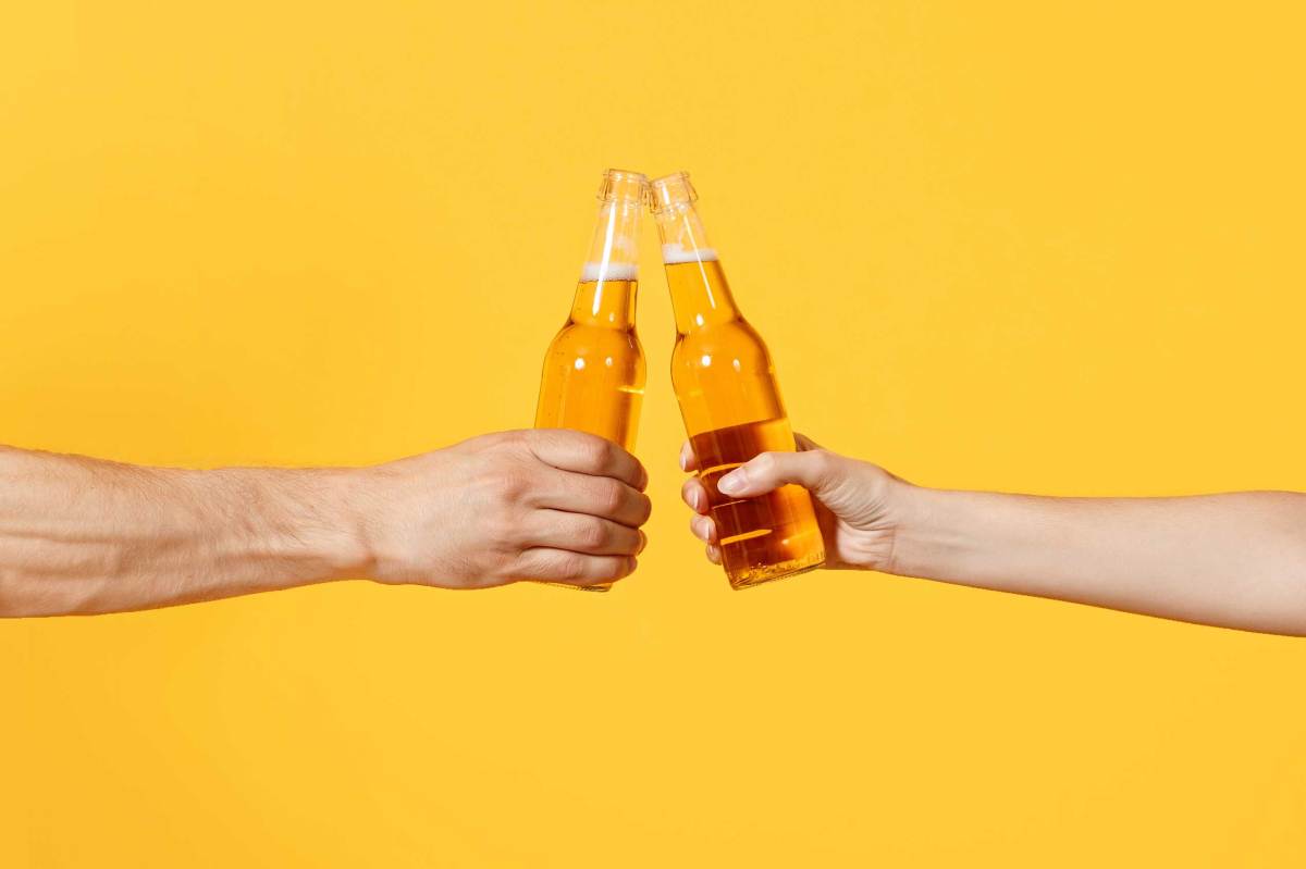Close up beschnitten von Frau und Mann zwei Hände horizontal halten Lagerbier Glasflaschen und stoßen an, isoliert auf gelbem Hintergrund.