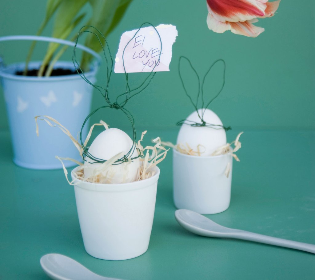 Hasen aus Draht auf Eiern in Eierbechern dahinter Blumenvase