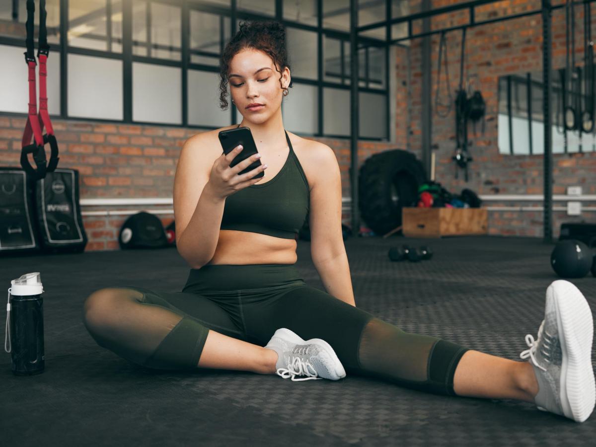 Frau im Fitnessstudio beim Workout, mit Smartphone und Fitness App. Junge Sportlerin mit Smartphone für Fortschritt, Leistung und Kommunikation für einen gesunden Lebensstil