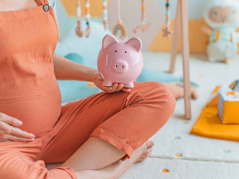 Schwangere Frau sitzt im Kinderzimmer und hält ein Sparschwein.