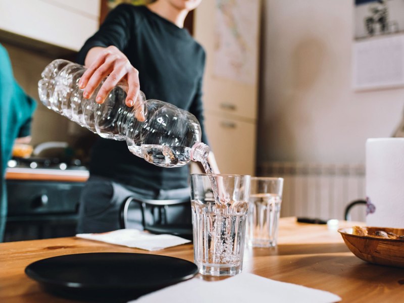 Frau schüttet Wasser aus Wasserflasche in ein Glas, dahinter ist eine Küche