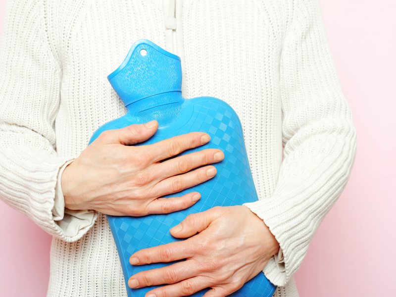 Hände umklammern eine blaue Wärmflasche vor weißem Strickpullover auf rosa Hintergrund.