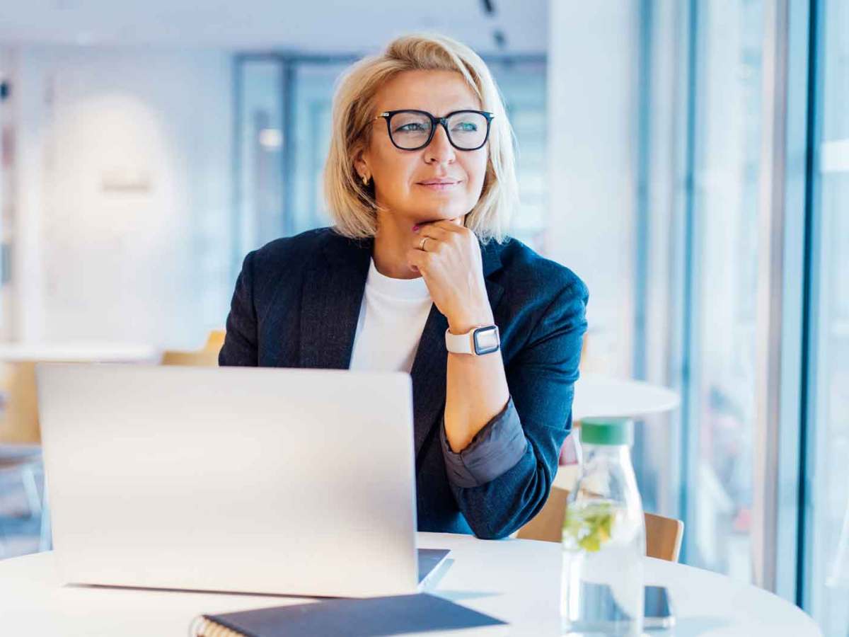 Businessfrau über 50 mit blonden Haaren und Brille sitzt im Büro an ihrem Laptop und denkt über ihren Urlaub nach.