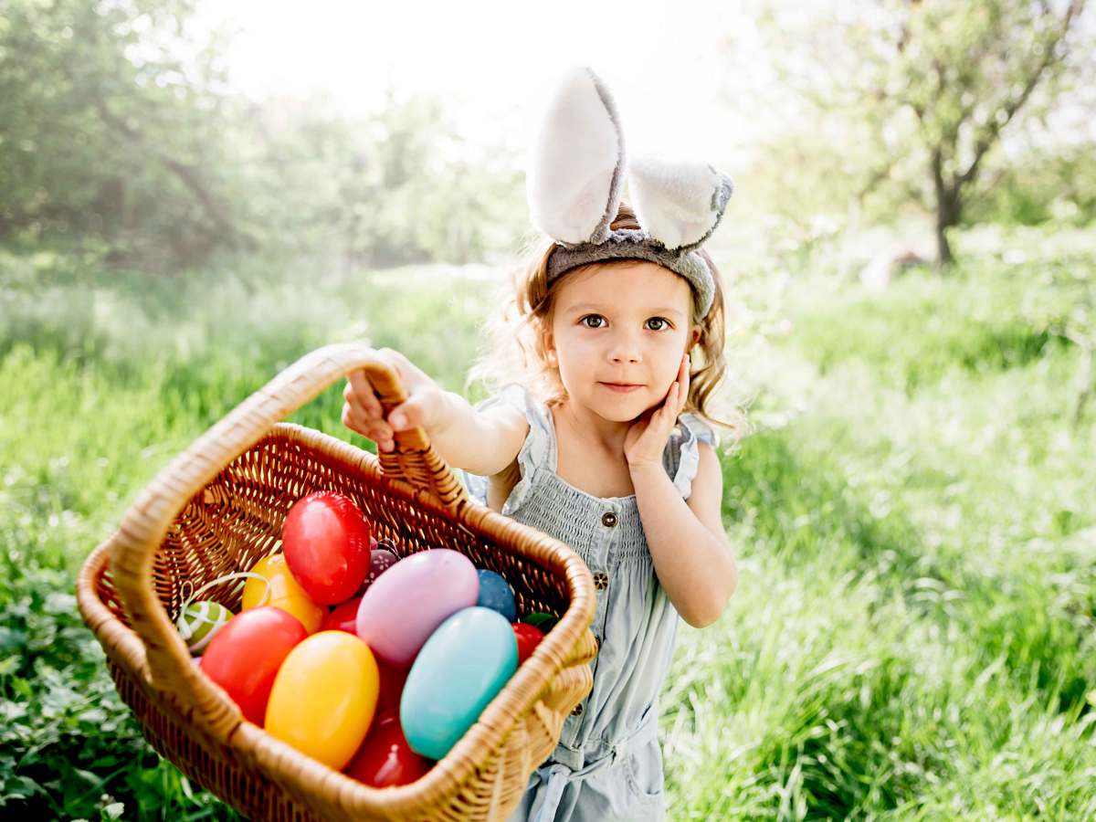 Kleines Mädchen mit Hasenohren auf dem Kopf zeigt stolz ihren prall gefüllten Osterkorb in die Kamera.