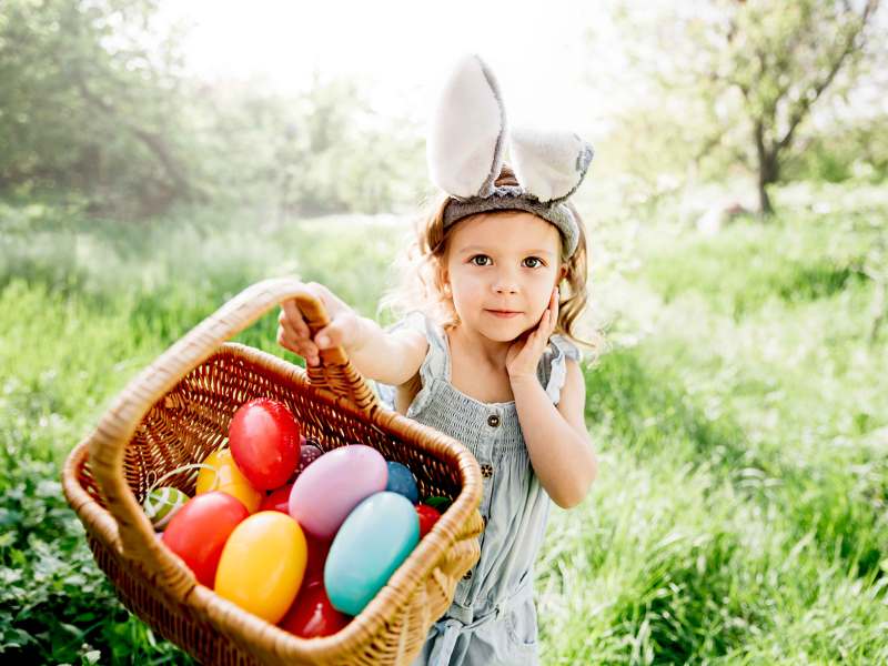 Kleines Mädchen mit Hasenohren auf dem Kopf zeigt stolz ihren prall gefüllten Osterkorb in die Kamera.
