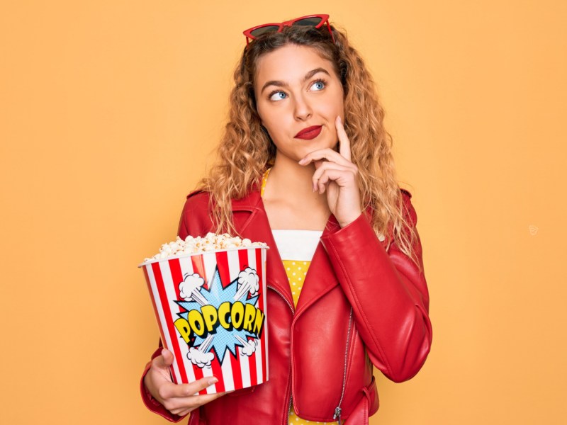 Frau vor gelber Wand mit Locken, die nachdenklich zur Seite schaut und ein Popcorn in der Hand hält