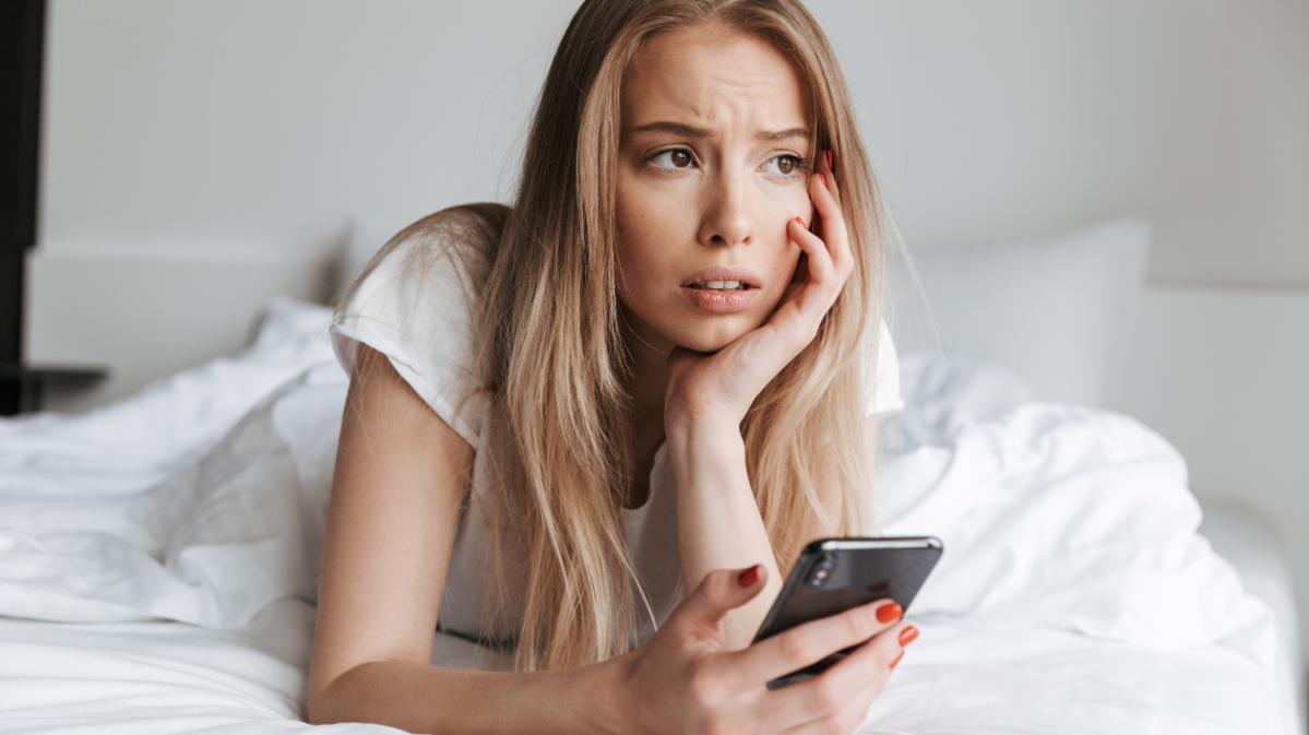 Eine junge Frau mit langen blonden Haaren liegt nachdenklich auf einem Bett, stützt ihren Kopf und hält ein Smartphone in der Hand.