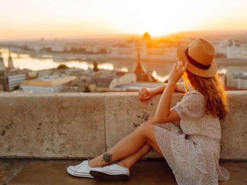 Frau mit braunen Haaren sitzt im Urlaub entspannt vor einer Urlaubskulisse und trägt ein Kleid und einen Hut.