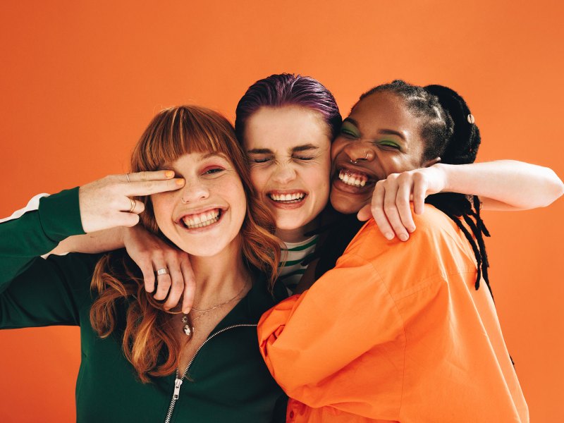 Drei Frauen umarmen sich vor einem orangefarbenen Hintergrund und lachen