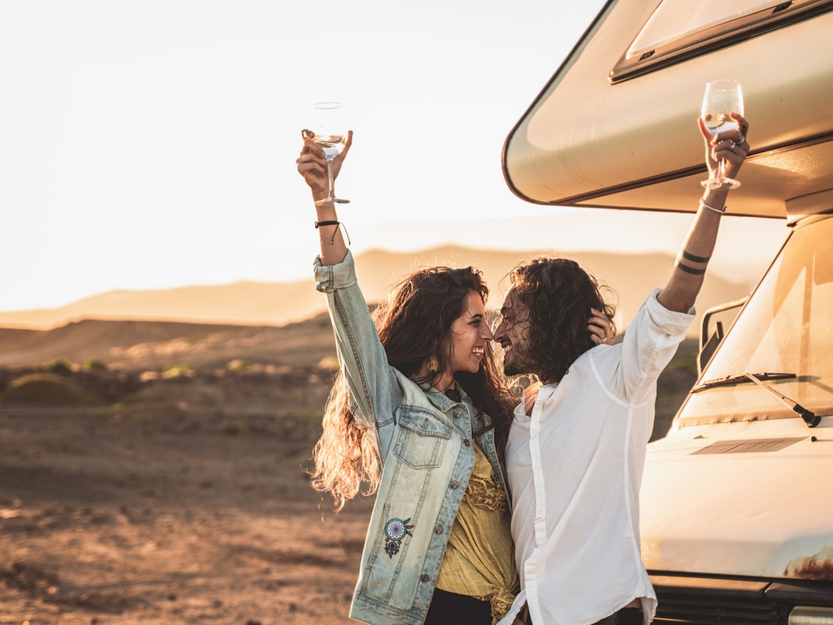 Ein Mann und eine Frau, die in einer Wüste stehen, ein Van im Hintergrund steht und beide ein Glas mit Wein in der Hand hochhalten und lächeln
