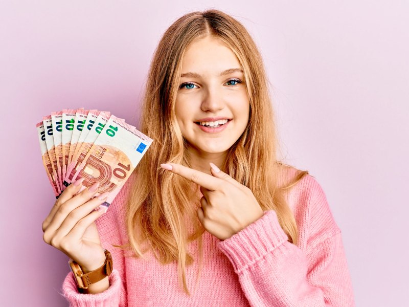 Mädchen vor rosa Hintergrund hält ein paar Zehn Euro Scheine in die Kamera und zeigt darauf.