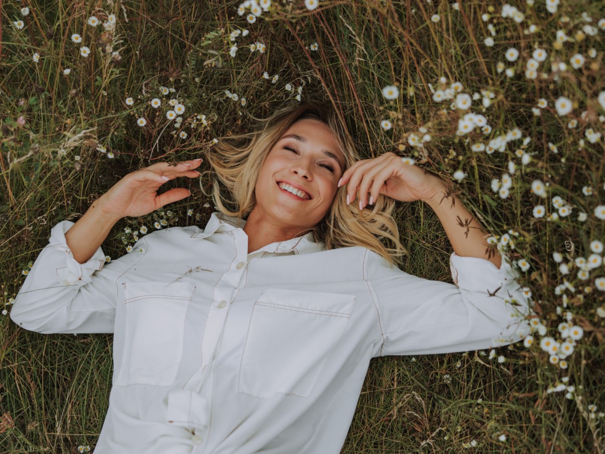 Frau auf einem Rasen mit Blumen, die eine weiße Bluse anhat und in die Kamera lächelt
