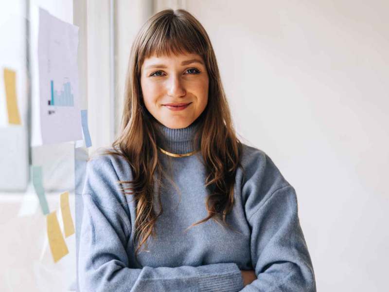 Braunhaarige Frau auf der Arbeit trägt einen hellblauen Pullover und eine goldene Kette. Sie steht mit verschränkten Armen und einem Lächeln vor einem Flipchart.