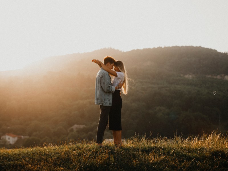 Frau und Mann auf einem Hügel einer Wiese, die sich küssen und von der Sonne bestrahlt werden