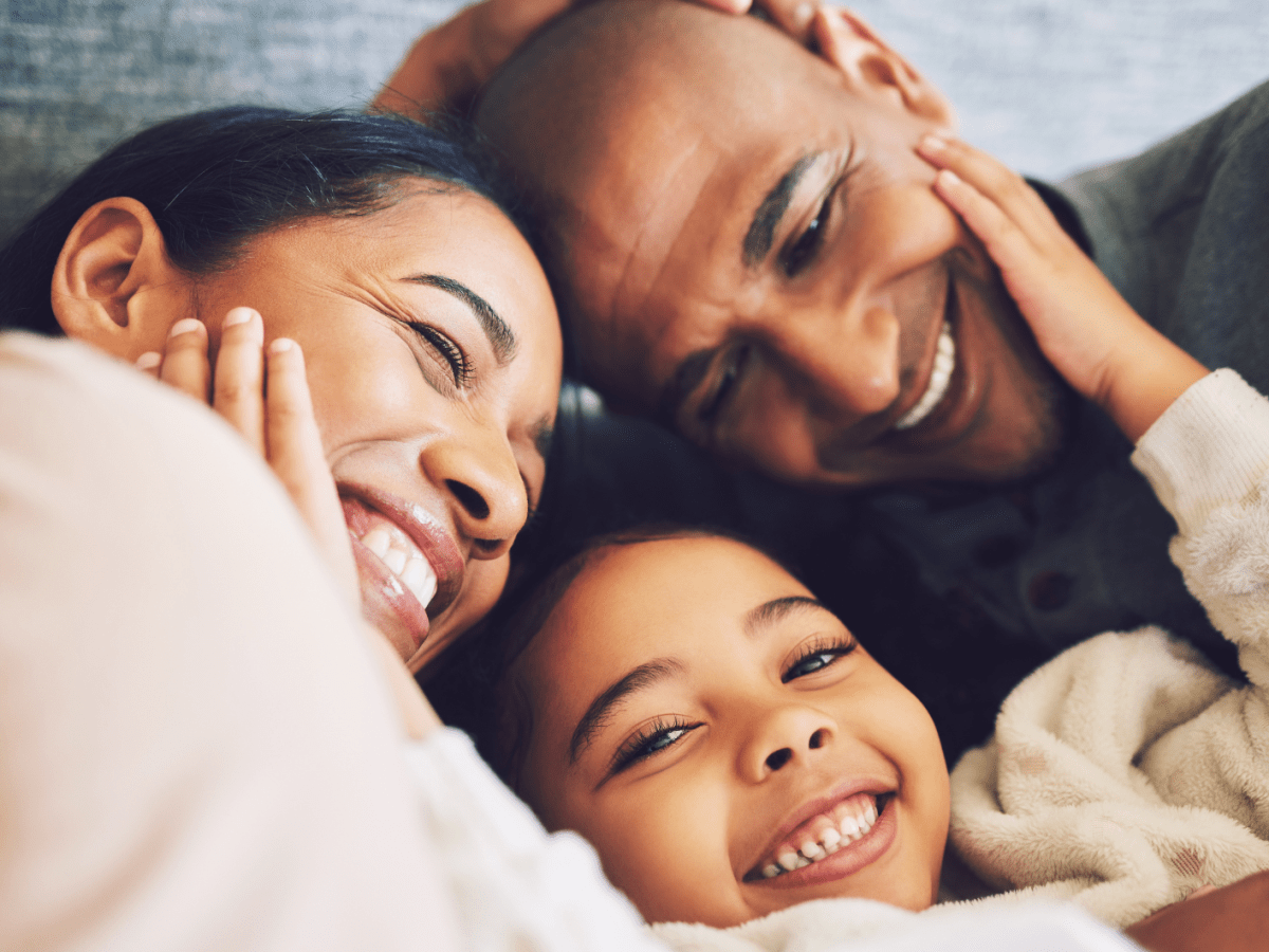 Familie, bestehend aus einer Mutter, einem Vater, einem Kind, die sich anlächelt und sich umarmt.