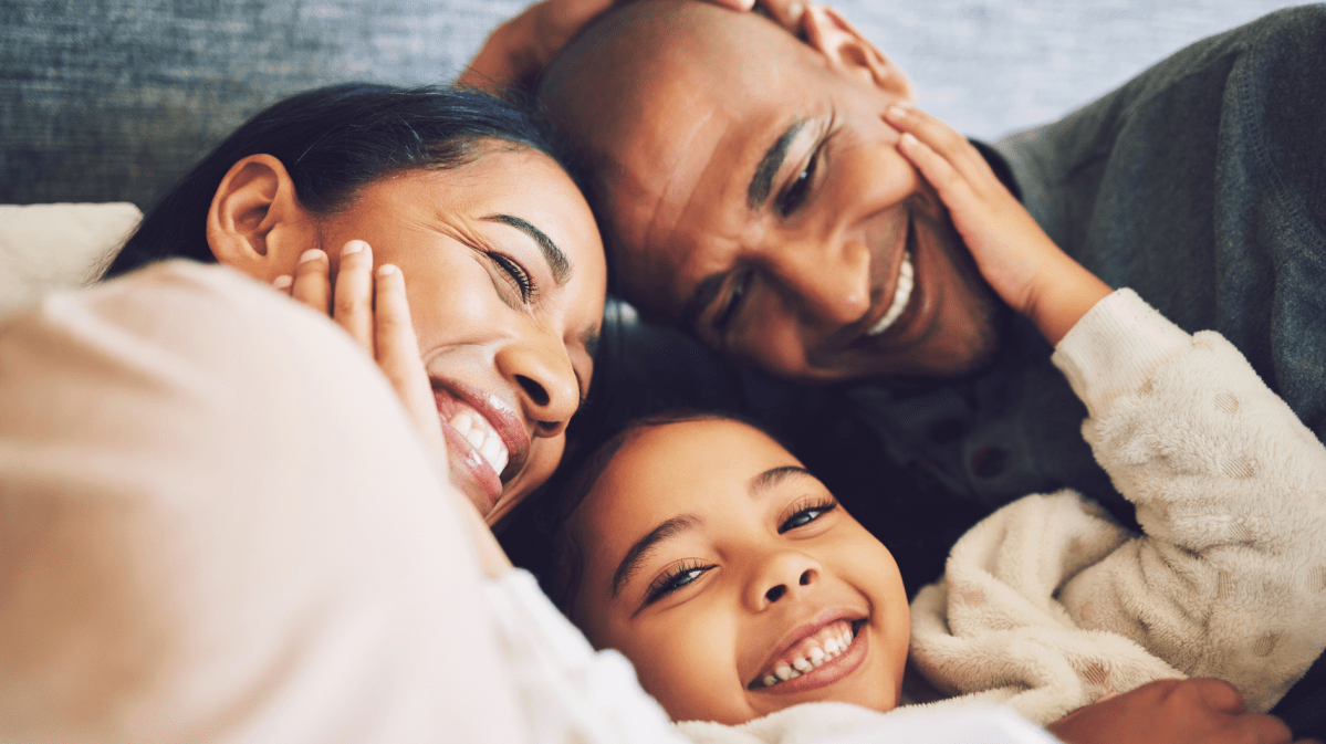 Familie, bestehend aus einer Mutter, einem Vater, einem Kind, die sich anlächelt und sich umarmt.