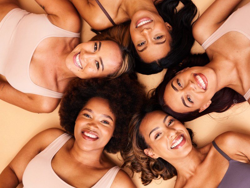 5 Frauen in einem Kreis, die auf dem Boden liegen, ihre Köpfe aneinander halten und in die Kamera lächeln.