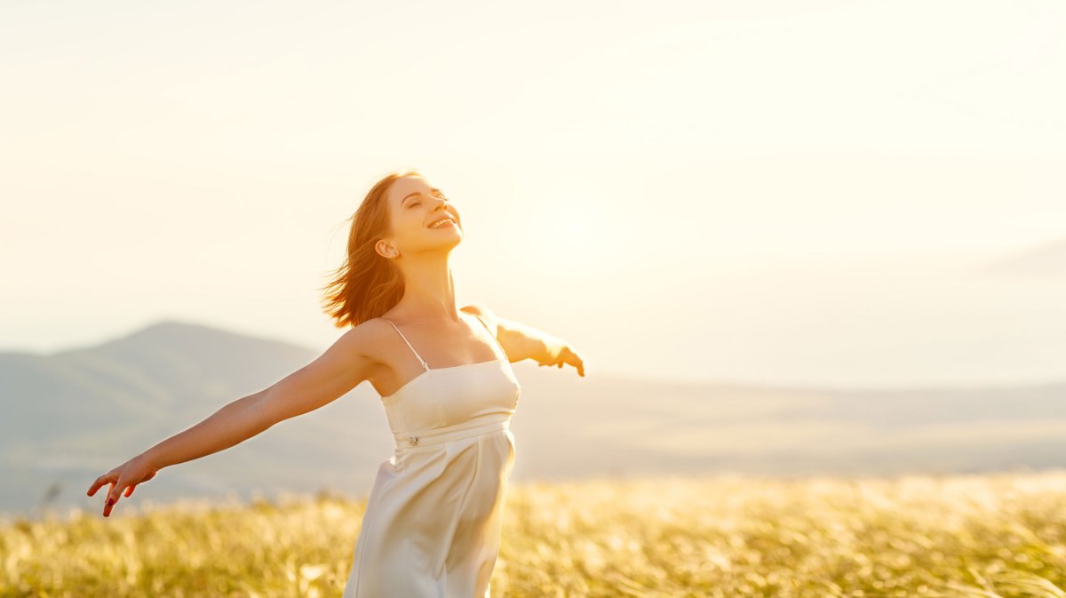 Frau in Sommerkleid mit ausgestreckten Armen auf einem Feld bei Sonnenschein.