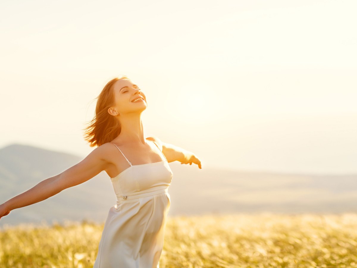 Frau in Sommerkleid mit ausgestreckten Armen auf einem Feld bei Sonnenschein.