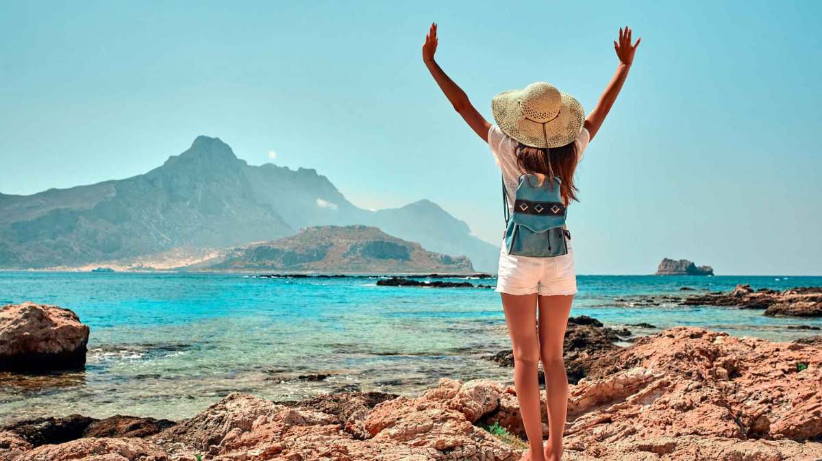 Junge Frau steht vor dem Meer auf einer Klippe und streckt die Arme nach oben. Sie trägt einen Sonnenhut, einen Rucksack und Sommerkleidung.