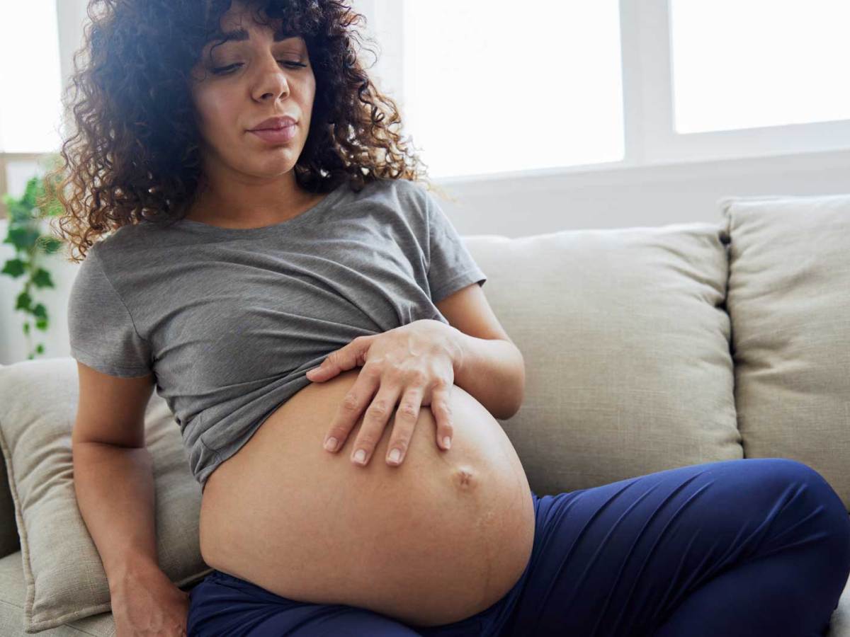 Schwangere Frau sitzt mit leicht schmerzverzerrtem Gesicht auf dem Sofa und hat eine Hand auf ihren Babybauch gelegt.