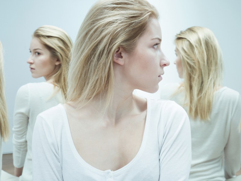 Blonde Frau sitzt vor mehreren Spiegeln und wird von Spiegelbildern angeschaut.