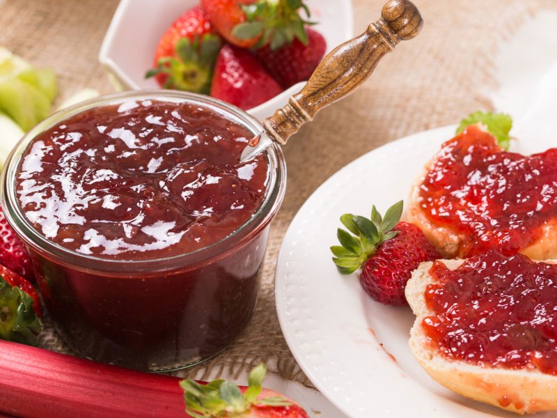 Fruchtaufstrich in Glas, neben weißem Teller mit zwei Brötchenhälften, die mit Marmelade beschmiert sind.