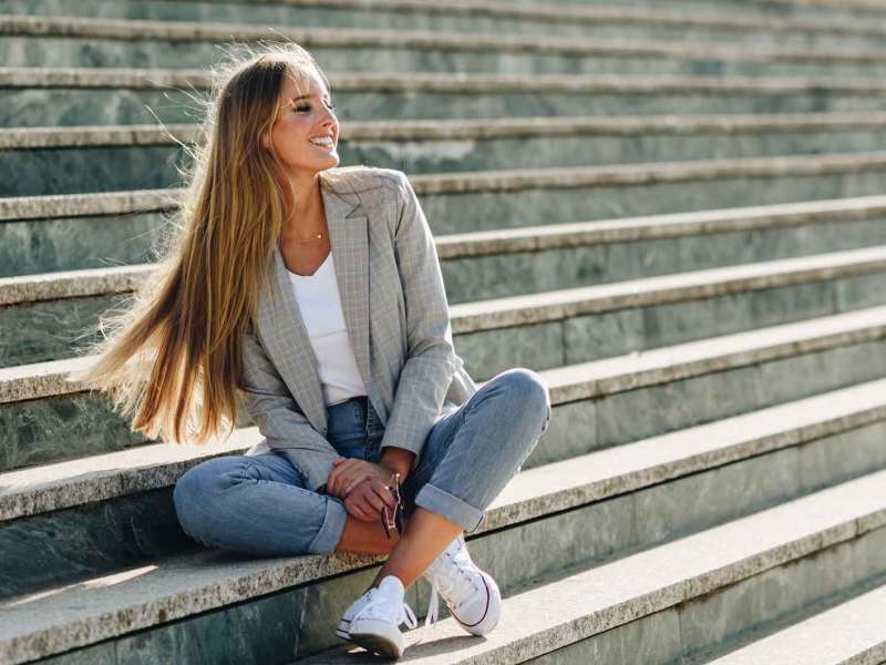 Frau mit langen Haaren sitzt draußen auf einer Treppe im Frühling und lacht.