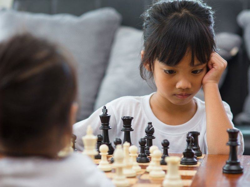 Zwei Mädchen, ca. 7 Jahre alt, spielen am Wohnzimmertisch eine Runde Schach. Ein Mädchen ist von hinten zu sehen, das andere von vorn. Es schaut konzentriert auf das Spielbrett.