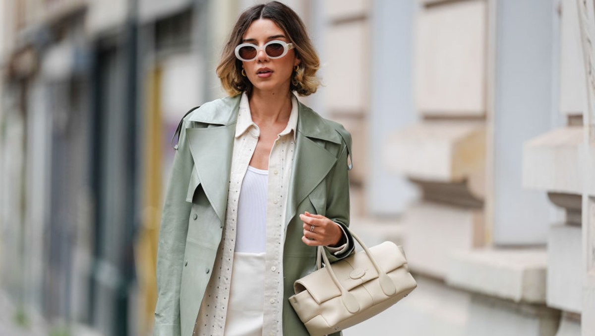 Katie Giorgadze wears sunglasses from Celine, golden earrings, a sage pastel green smooth leather trench coat from Mackage, a cream colored mini skirt, a white tank top from Uniqlo, a beige oversize rhinestone sparkly leather shirt from Frame, a cream color / white leather bag from Tod's, during a street style fashion photo session, on April 07, 2024 in Paris, France.