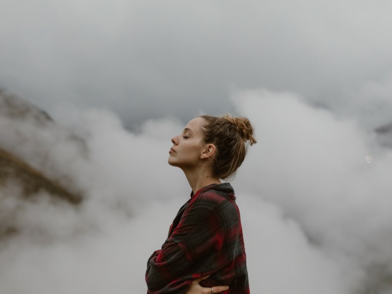 Frau auf einem Berg mit Wolken im Hintergrund, die ihre Augen schließt und einatmet