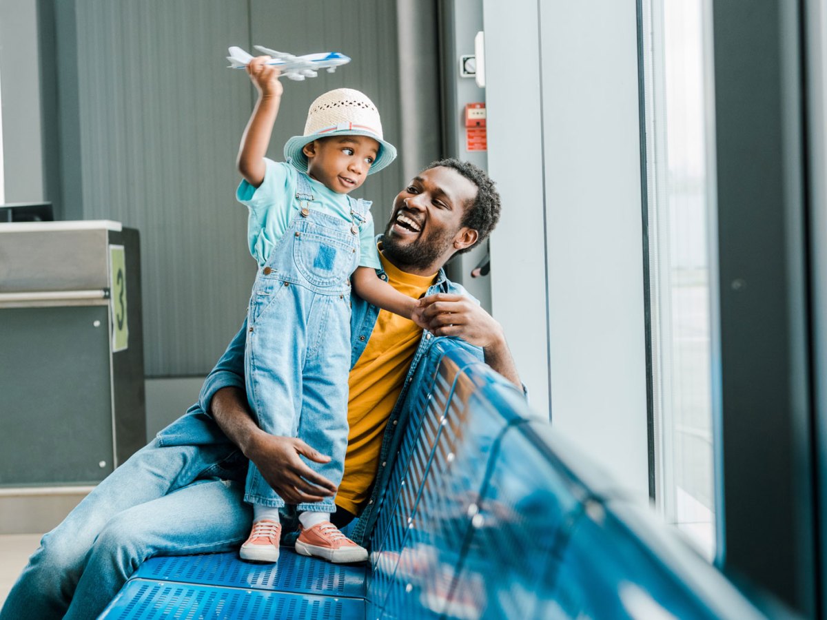 Vater und Sohn am Flughafen. Beide sind am Fenster, der Sohn steht auf dem Sitz und spielt mit einem Spielzeugflugzeug.
