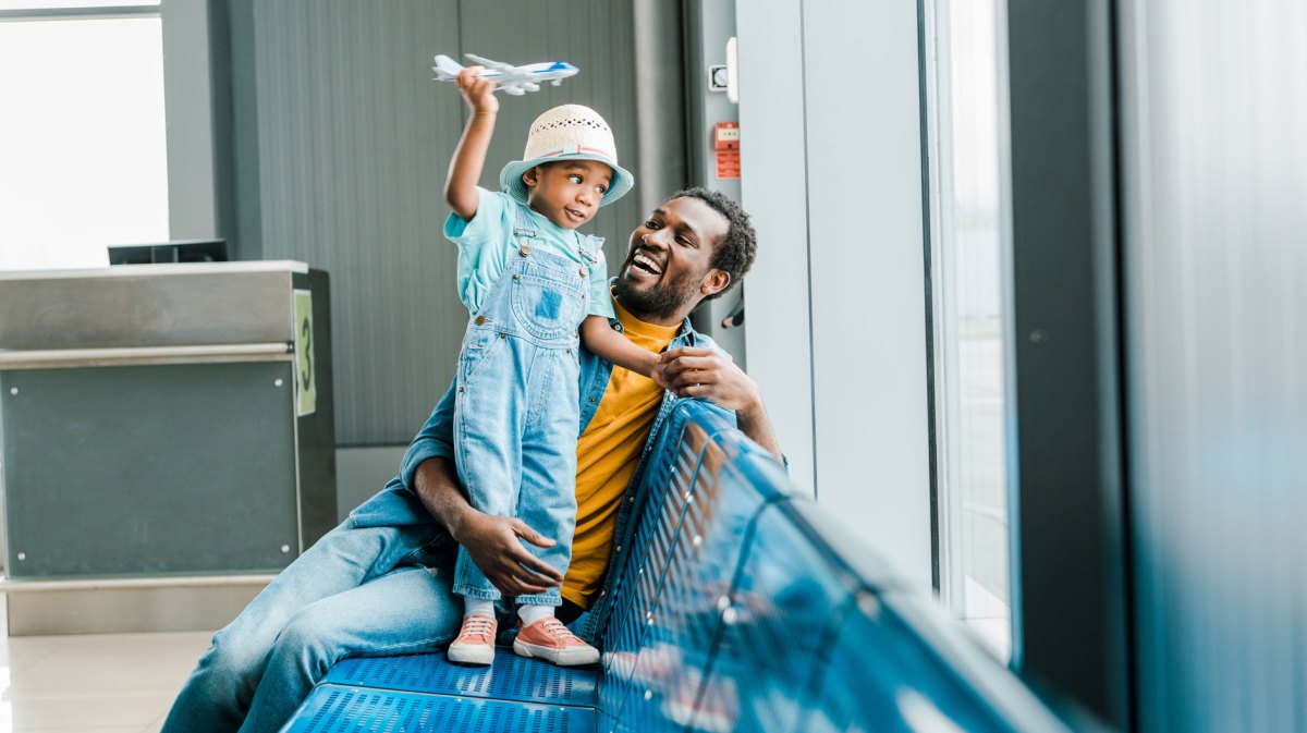 Vater und Sohn am Flughafen. Beide sind am Fenster, der Sohn steht auf dem Sitz und spielt mit einem Spielzeugflugzeug.