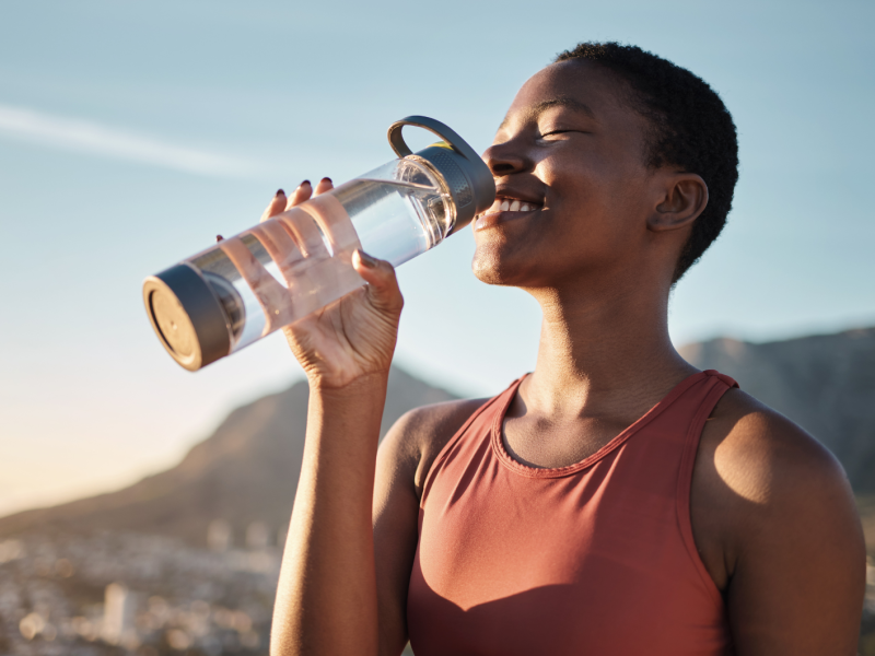 Frau auf einem Berg mit Landschaft im Hintergrund, die glücklich aus einer Wasserflasche trinkt.