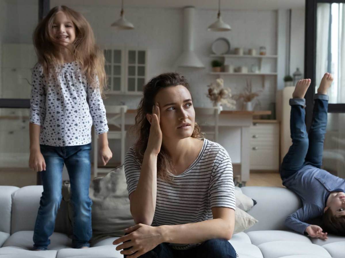 Mutter sitzt genervt auf dem Sofa, während ihre beiden Kinder um sie herum turnen.