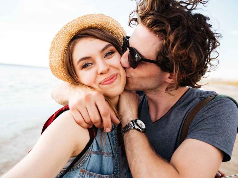 Junges Paar am Strand im Urlaub hat Spaß. Er küsst sie auf die Wange und sie lächelt in die Kamera.