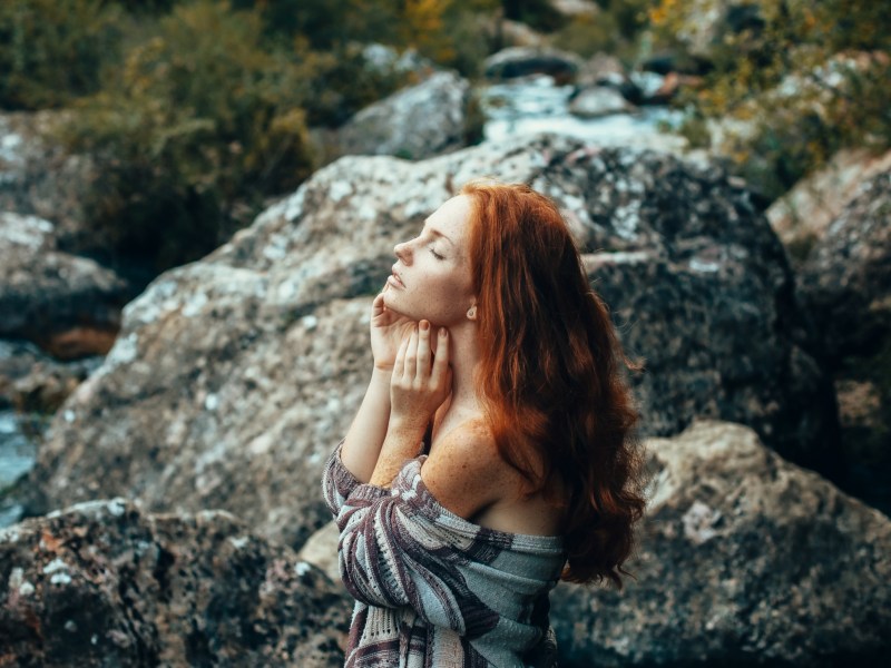 Frau in der Natur vor einem Bach mit Steinen, die ihre Augen schließt und ihre Hände auf dem Kinn abstützt