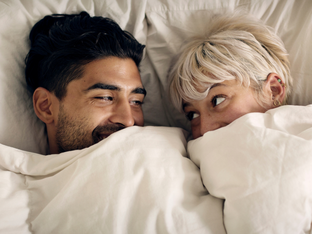 Ein dunkelhaariger Mann und eine blondhaarige Frau schauen sich lächelnd unter einer weißen Bettdecke verliebt in die Augen und lachen.