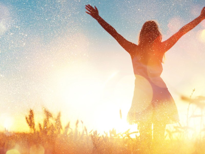 Frau im Sommerkleid mit ausgestreckten Armen auf einem Feld bei Sonnenuntergang