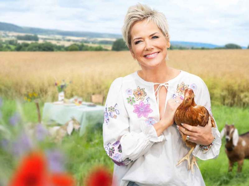 Inka Bause steht auf einem Feld und hält ein Huhn im Arm.