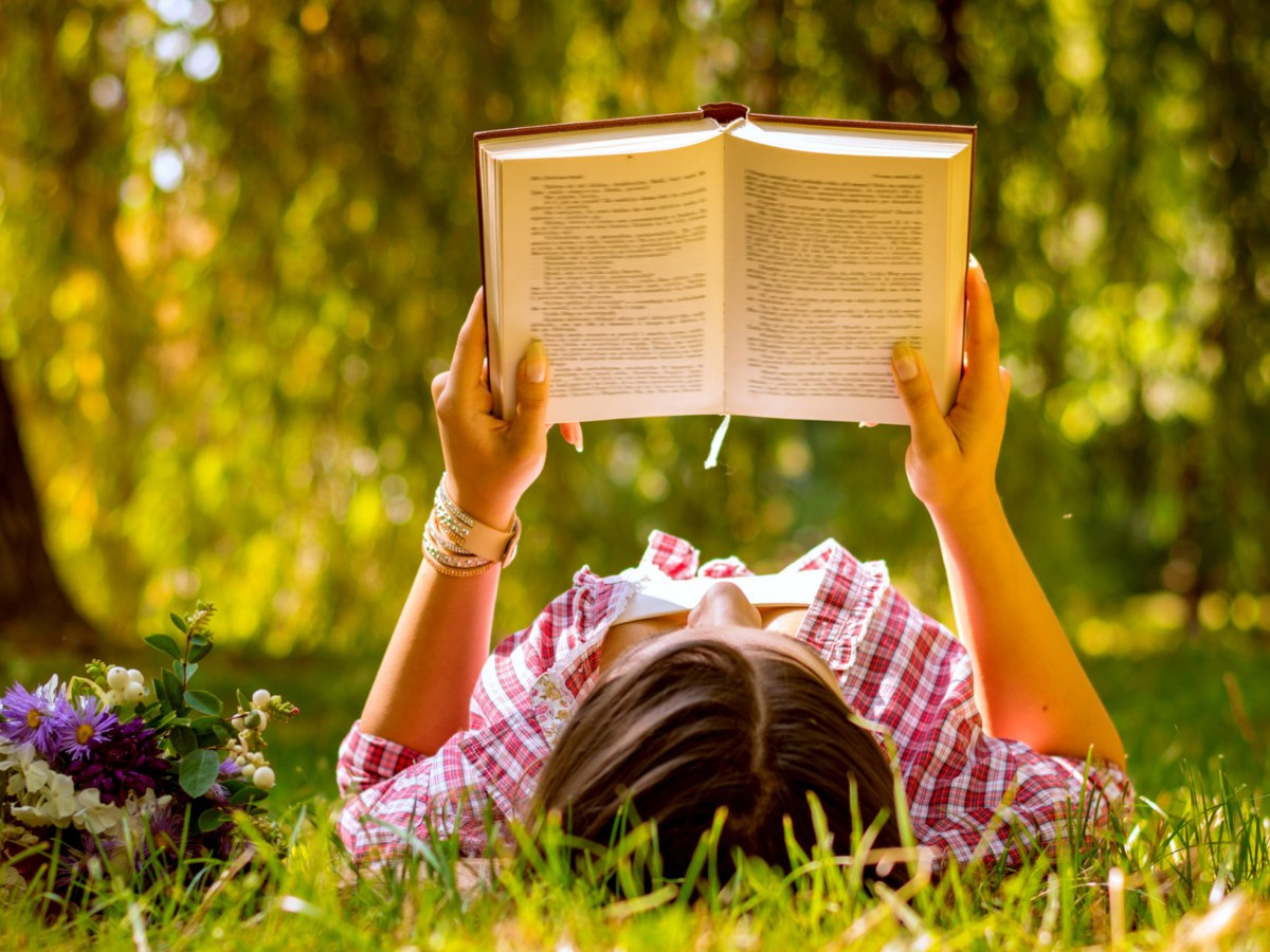 Frau liegt auf Wiese und liest Buch.