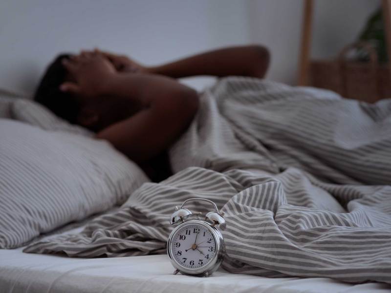 Müde Person hält sich im Bett mit gestreifter Bettwäsche die Hände vor das Gesicht, Wecker im Vordergrund.